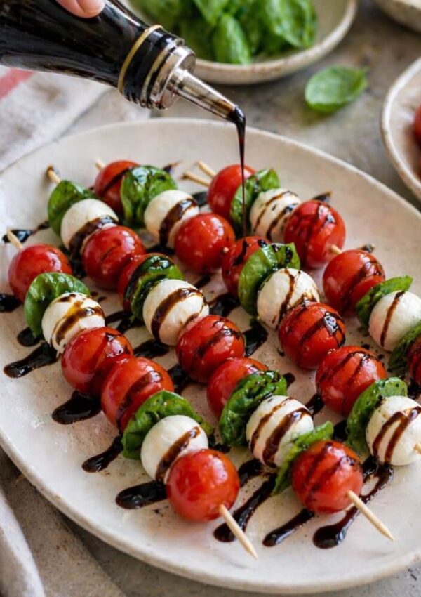 A hand drizzles balsamic glaze over skewers of cherry tomatoes, mozzarella balls, and fresh basil leaves, arranged neatly on a white platter.