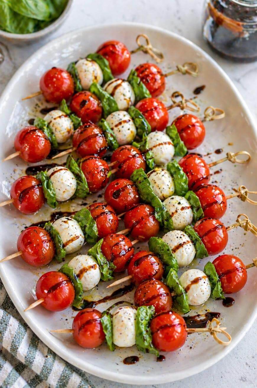 A platter of Caprese skewers with cherry tomatoes, mozzarella balls, and fresh basil leaves, drizzled with balsamic glaze, arranged on a white plate.