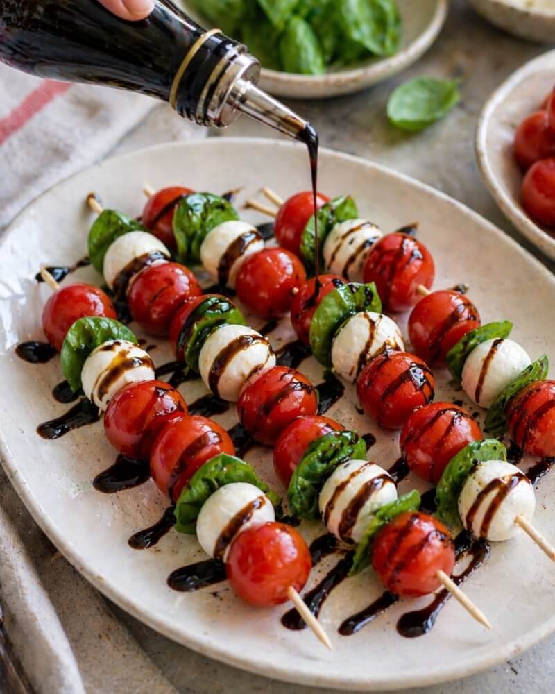 A hand drizzles balsamic glaze over skewers of cherry tomatoes, mozzarella balls, and fresh basil leaves, arranged neatly on a white platter.