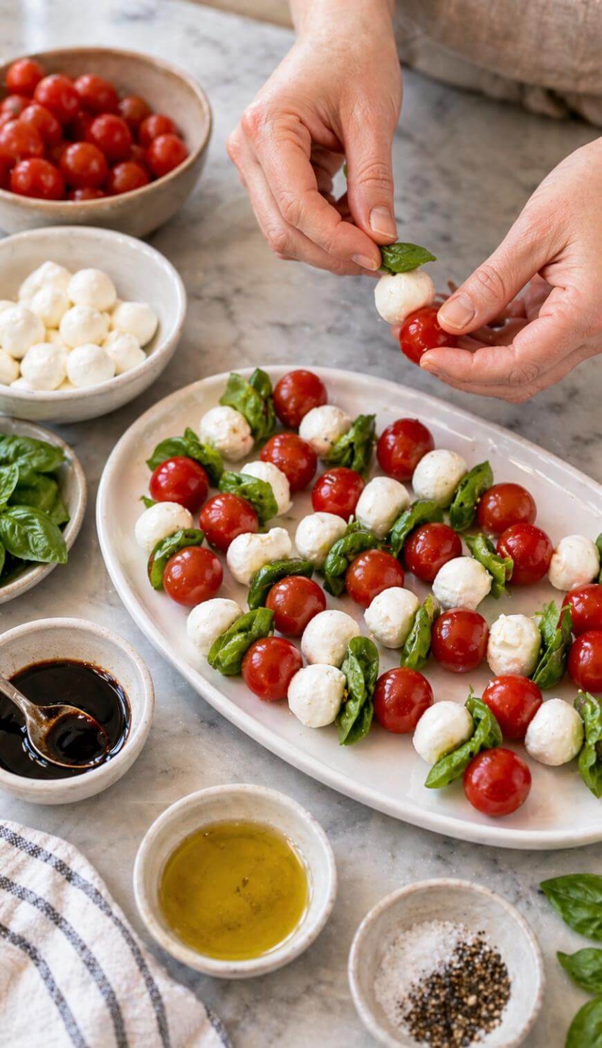 Hands assembling Caprese skewers with cherry tomatoes, mozzarella balls, and basil leaves on a white platter. Bowls of tomatoes, mozzarella, basil, balsamic glaze, olive oil, and pepper are on the marble countertop.