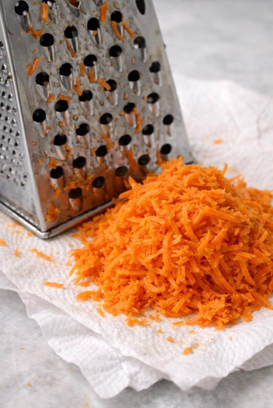A metal box grater sits on a paper towel next to a pile of freshly grated carrots on a light surface.