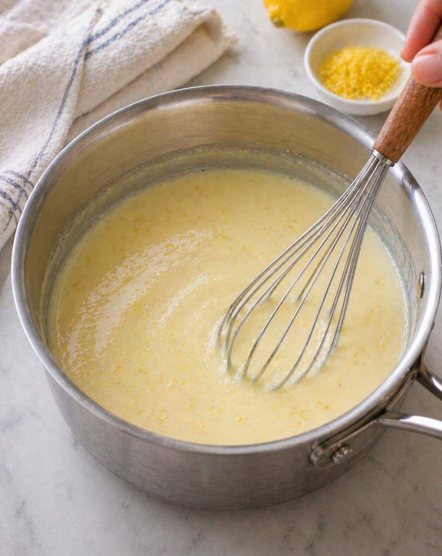 A hand holding a whisk stirs creamy yellow lemon mixture in a stainless steel saucepan on a marble countertop. A kitchen towel, a lemon, and a small bowl of lemon zest are nearby.