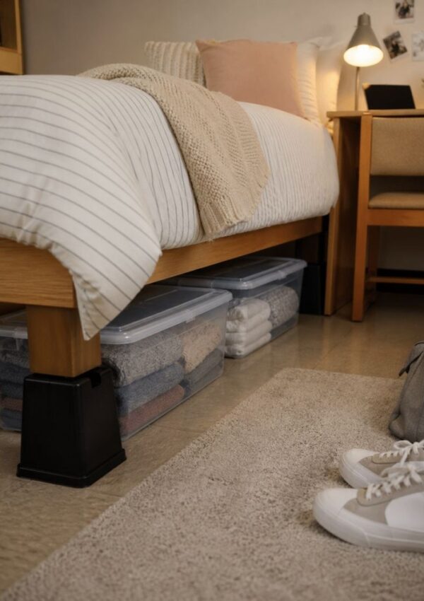 A neatly made bed on risers with storage bins of folded clothes underneath—perfect for storing freshman year essentials—a beige rug, white sneakers, and a desk with a lamp and plants complete this cozy dorm setup.
