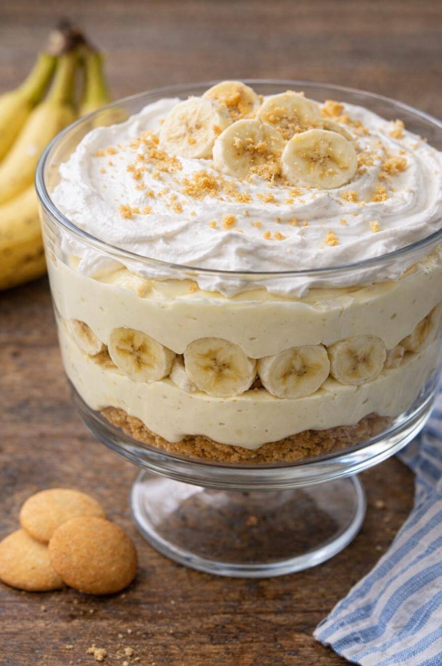 A glass trifle dish filled with layered banana pudding, sliced bananas, whipped cream, and sprinkled cookie crumbs, with whole bananas and vanilla wafers in the background on a wooden table.