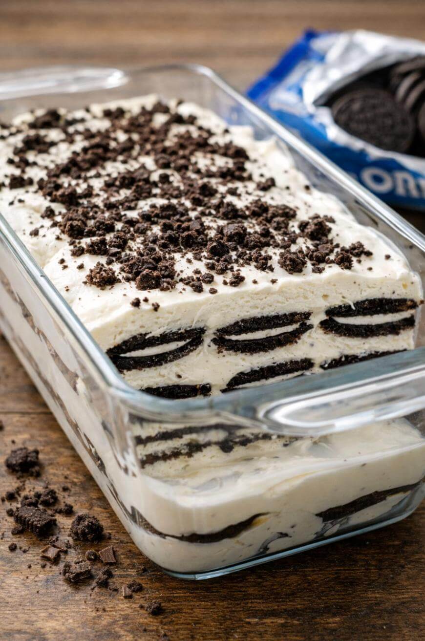 A glass dish filled with layers of whipped cream and whole Oreo cookies, topped with crumbled Oreos, sits on a wooden table with an open package of Oreo cookies in the background.