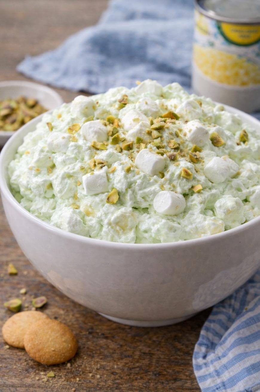 A white bowl filled with green pistachio fluff salad, topped with mini marshmallows and chopped pistachios, sits on a wooden table with cookies and a can of fruit in the background.
