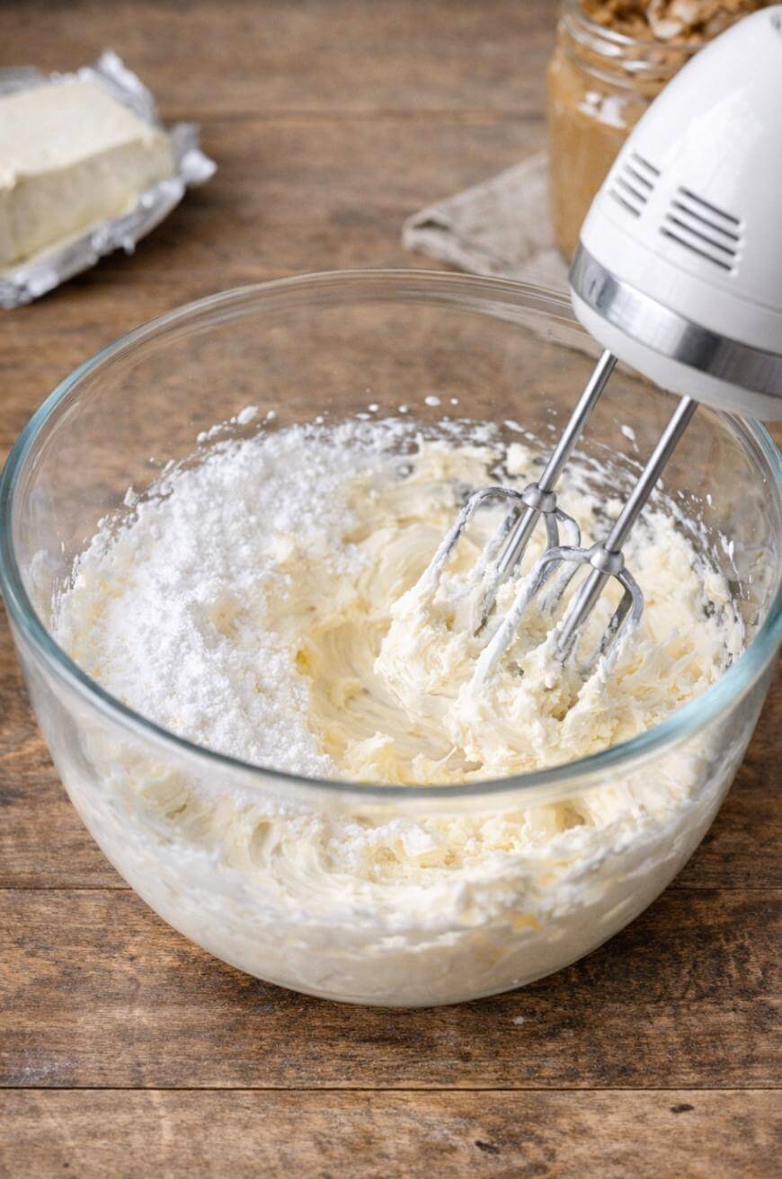 A hand mixer blends powdered sugar and cream cheese in a glass bowl on a wooden surface, with a block of cream cheese and a jar of peanut butter in the background.