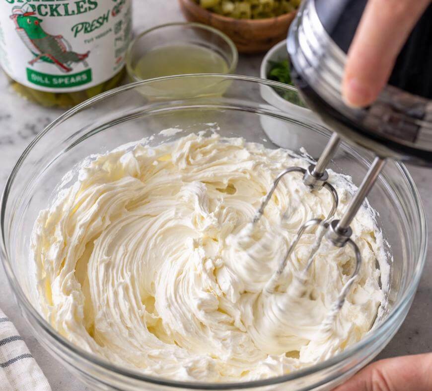 A hand holds an electric mixer blending creamy white ingredients in a glass bowl. Nearby are a jar of pickles, a small bowl of chopped pickles, and a dish with liquid on a marble countertop.