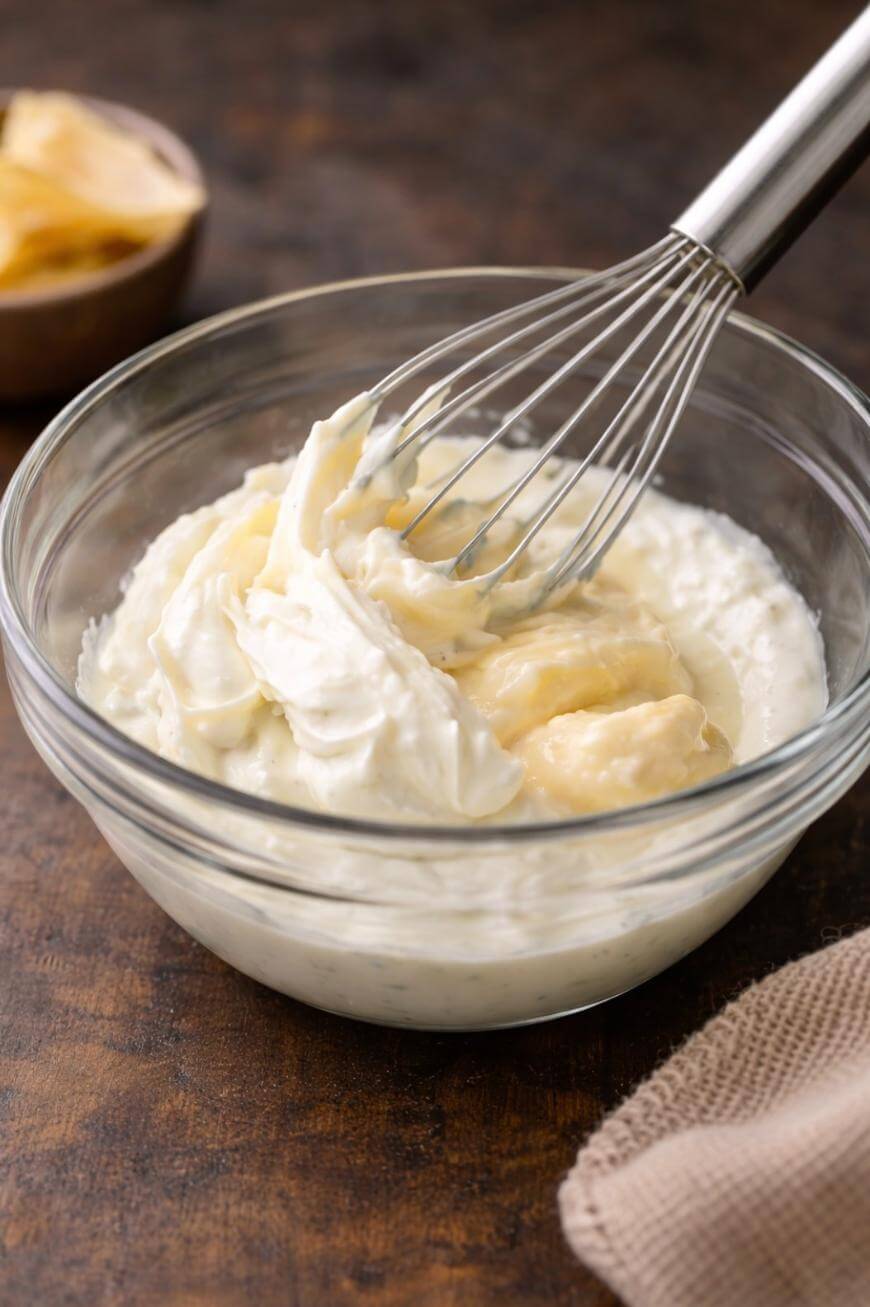 A glass bowl with mashed bananas and creamy yogurt being mixed with a metal whisk, set on a wooden surface with a beige napkin nearby.