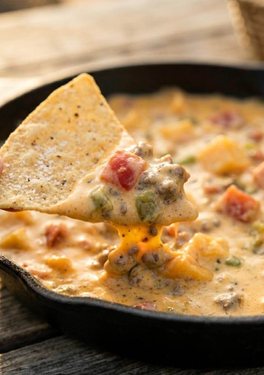 A close-up of a tortilla chip scooping creamy queso dip with ground beef, tomatoes, and green peppers from a cast iron skillet.