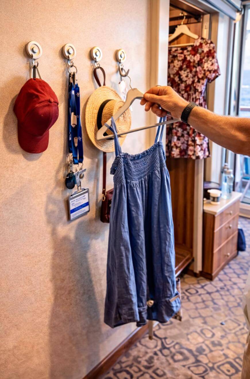 A hand holds a blue sundress on a hanger in a room with hats, a name badge, a lanyard, and a red cap hanging on hooks on the wall. A floral dress hangs in an open closet nearby.