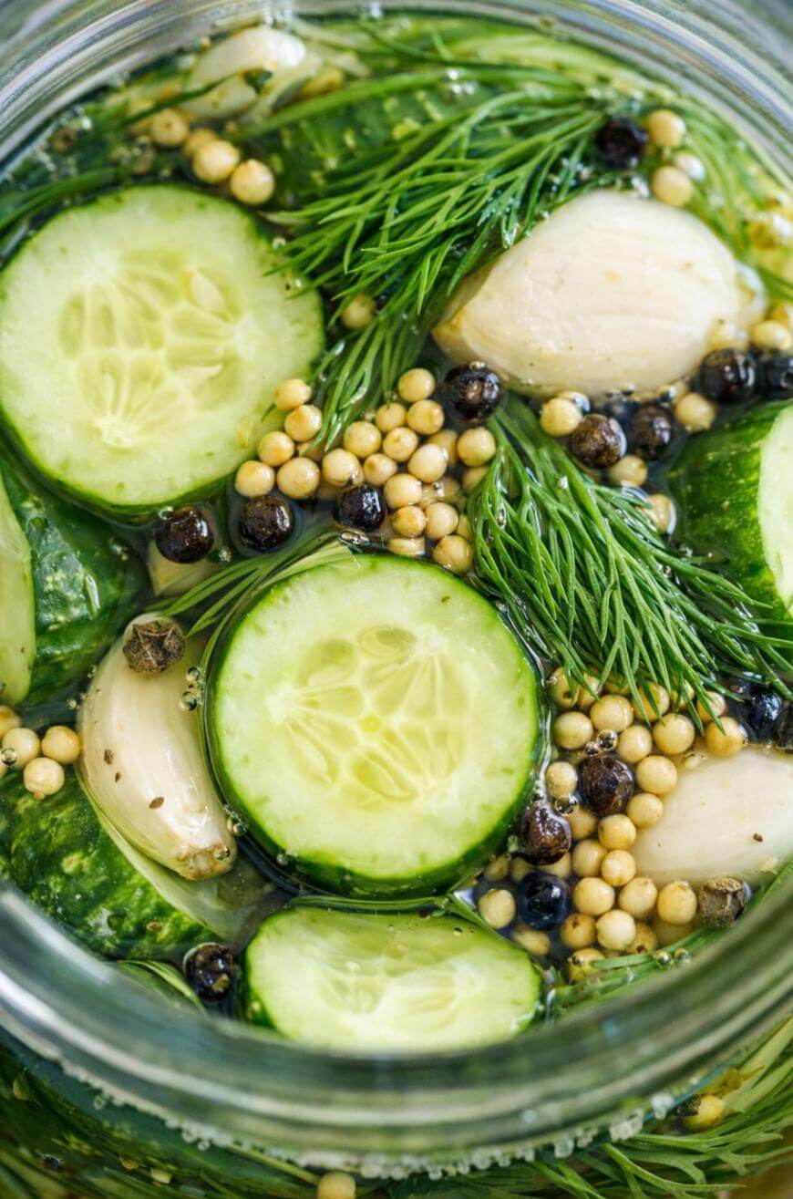 Sliced cucumbers, garlic cloves, fresh dill, mustard seeds, and black peppercorns submerged in brine, seen from above in a glass jar, as ingredients for homemade pickles.