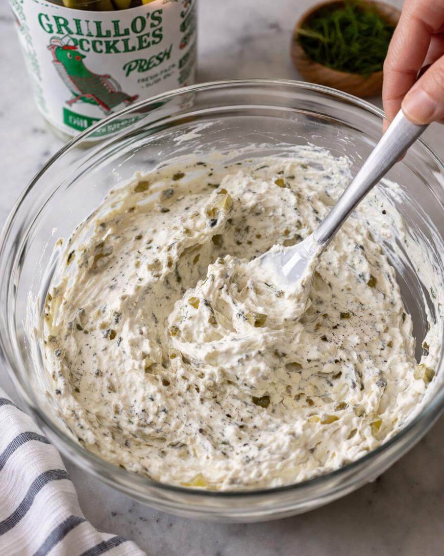 A hand stirs a creamy mixture with chopped pickles in a glass bowl, using a white spatula. An open container of Grillo’s Pickles and a small bowl of herbs are on the countertop nearby.