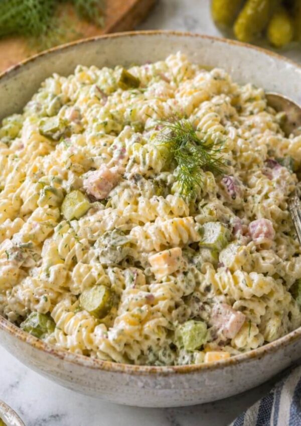 A large bowl of creamy pasta salad with rotini noodles, diced pickles, ham, cheese cubes, and fresh dill, with a silver serving spoon resting in the bowl.