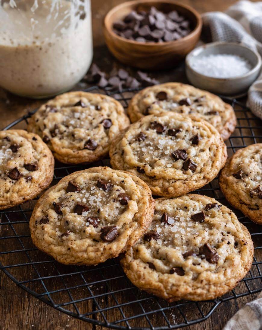 A batch of chocolate chip cookies sprinkled with flaky sea salt cooling on a wire rack, with a jar of dough, a bowl of chocolate chips, and a bowl of salt in the background.