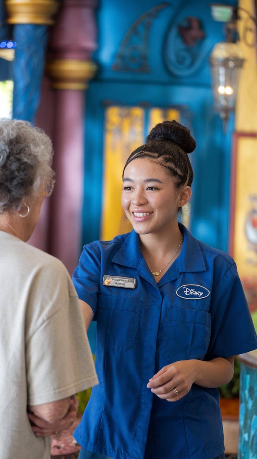 A young woman in a blue Disney uniform smiles warmly while greeting an older woman with gray hair in a beige shirt inside a brightly decorated indoor space.