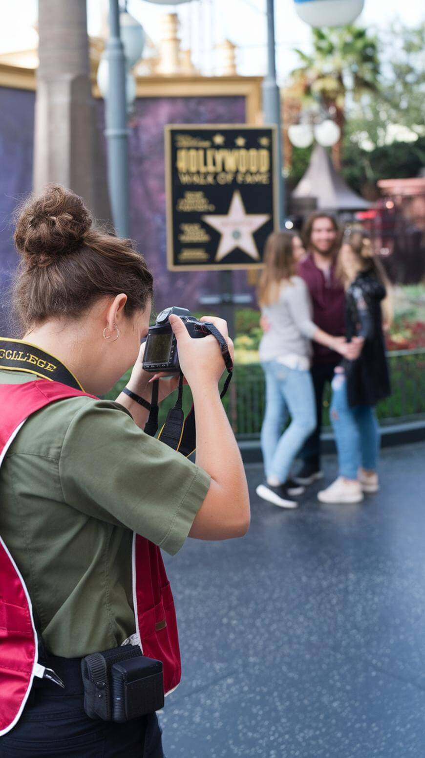 A photographer wearing a red vest takes a picture of three people posing together in front of a Hollywood-themed sign outdoors. The group stands on a sidewalk, smiling and holding hands.