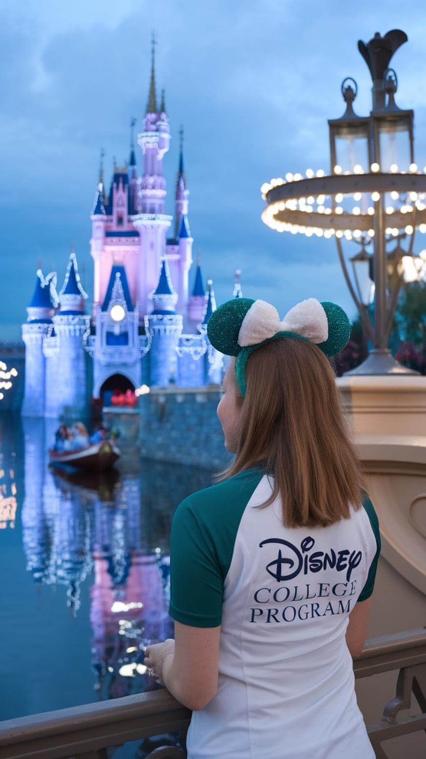 A woman wearing Mickey ears and a Disney College Program shirt stands by a railing, looking at a brightly lit Disney castle across the water at dusk. A boat floats on the water in the background.