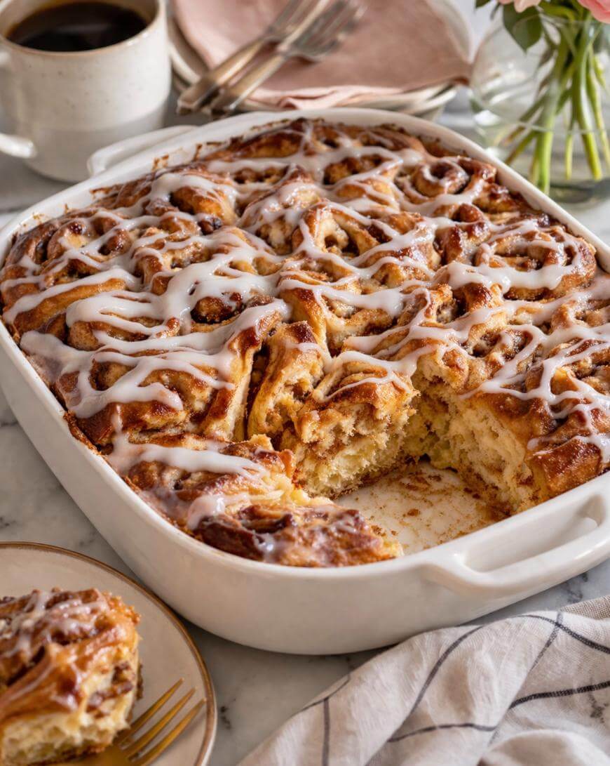 A baking dish filled with iced cinnamon rolls, one piece missing, sits on a table. The rolls are drizzled with white icing, and a single piece is on a plate nearby. A cup of coffee, utensils, and a napkin are also visible.