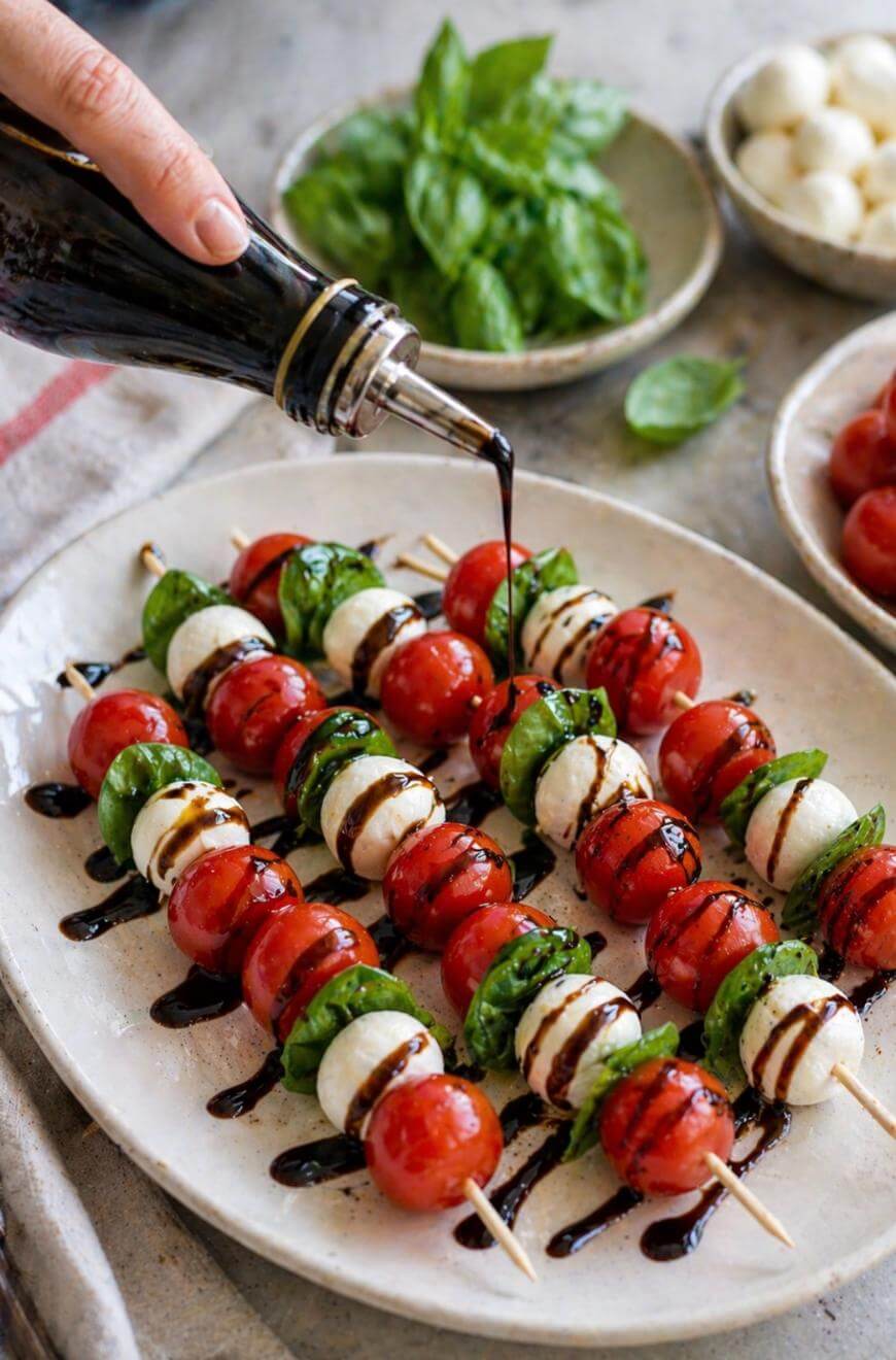 A hand drizzles balsamic glaze over skewers with cherry tomatoes, mozzarella balls, and fresh basil leaves on a white plate, with bowls of basil and cheese in the background.