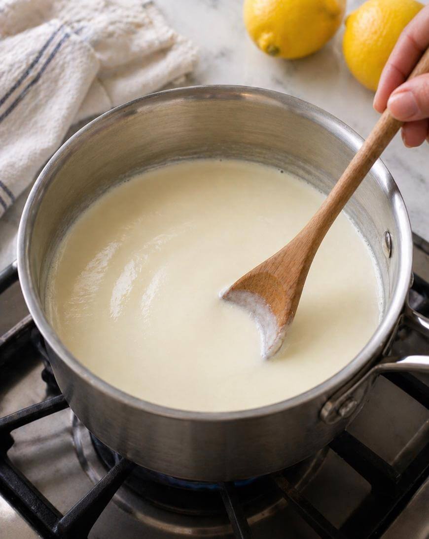 A hand stirs a creamy white sauce in a saucepan with a wooden spoon on a stovetop; lemons and a kitchen towel are in the background.