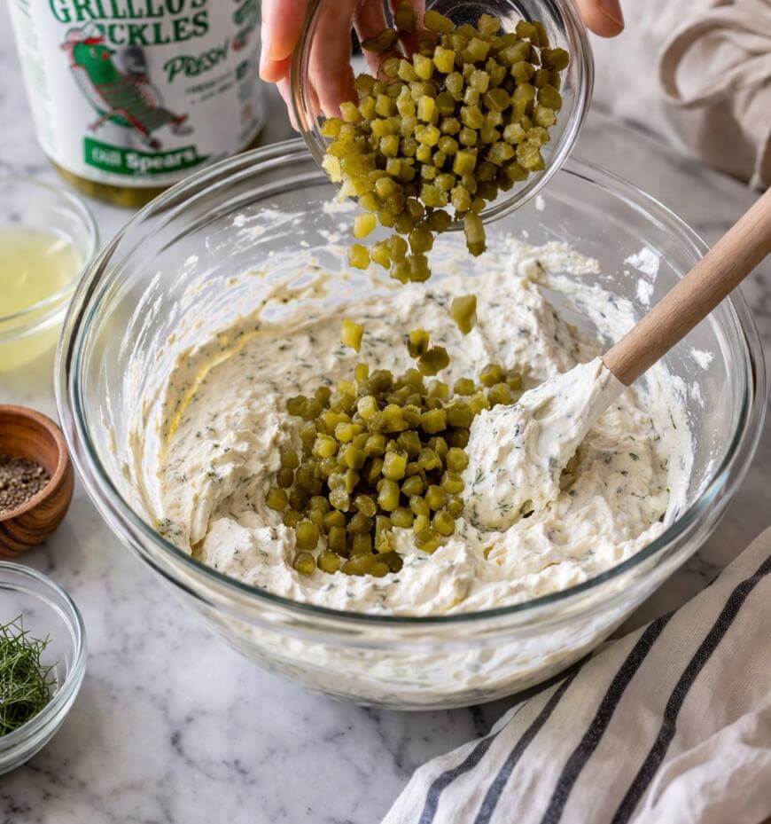 A hand pours diced pickles from a small bowl into a larger glass bowl filled with a creamy mixture, with a wooden spoon inside. Other ingredients and a jar of pickles are nearby on a marble countertop.