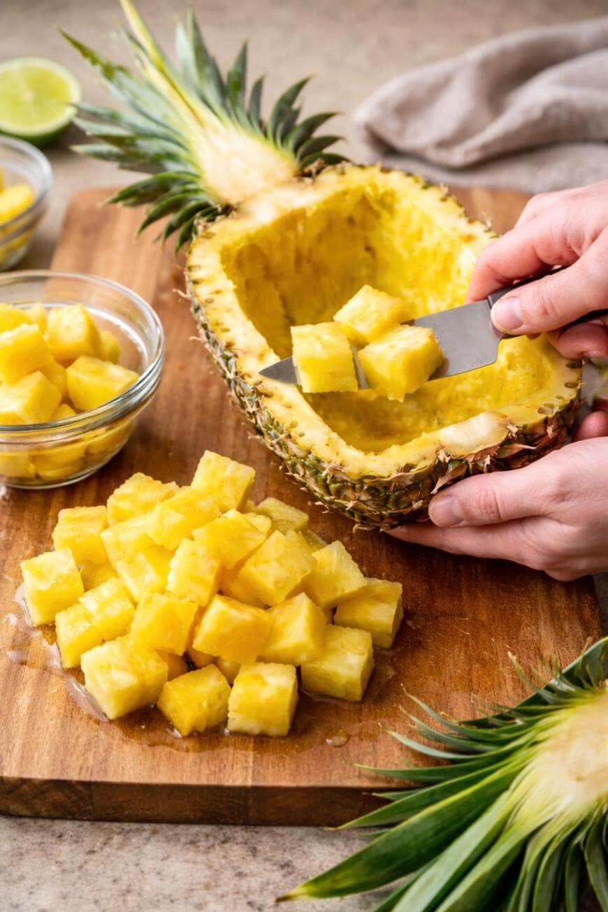 A person uses a knife to cut pineapple cubes, placing them into a hollowed-out pineapple half on a wooden cutting board. Bowls and piles of pineapple cubes surround the board.