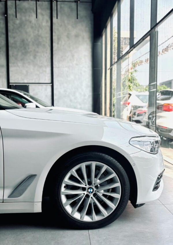 Side view of a white BMW car parked indoors near large glass windows, with sunlight streaming in and other cars visible outside.