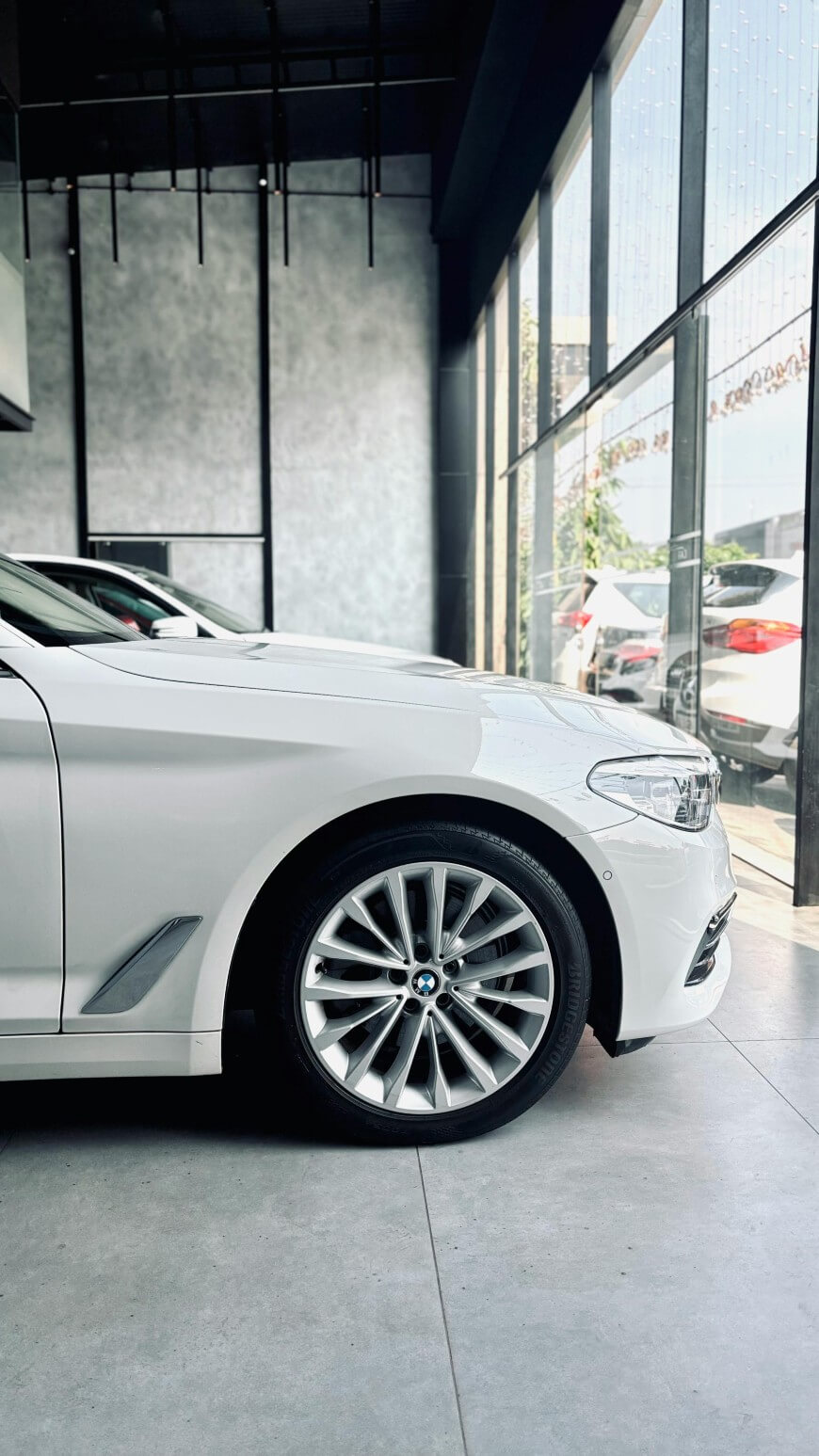A white BMW sedan is parked indoors near large glass windows; sunlight shines in, reflecting on the cars body and alloy wheel. Other cars are visible outside through the glass.