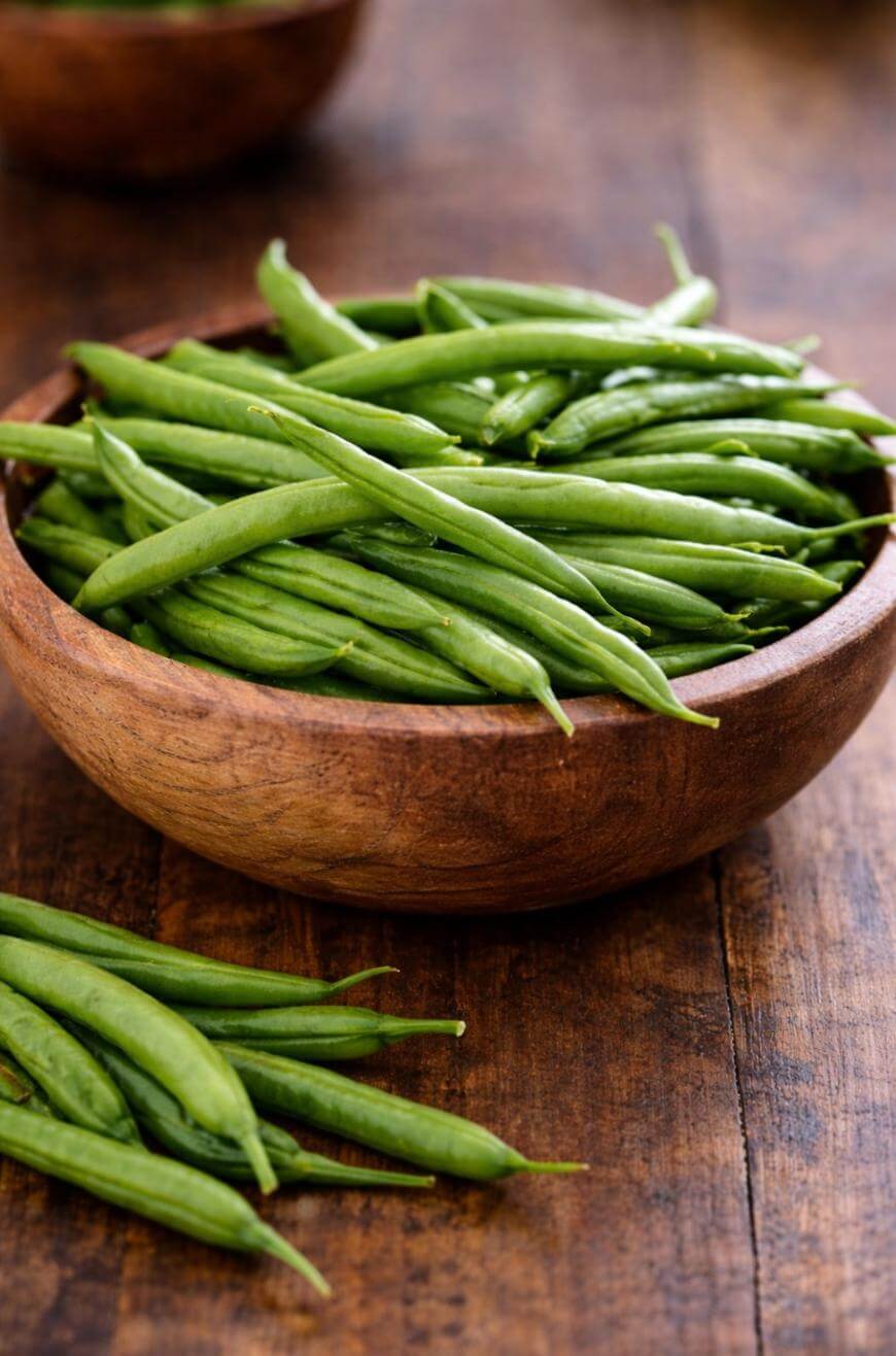 A wooden bowl filled with fresh green beans sits on a rustic wooden table, with a few green beans scattered in front of the bowl.