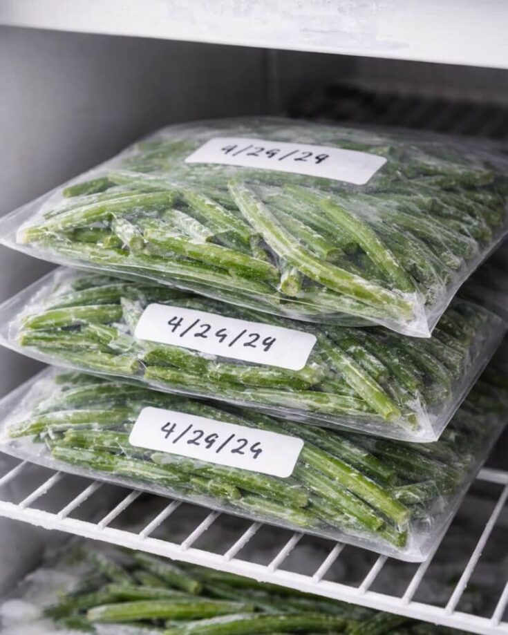 Three sealed plastic bags filled with frozen green beans are stacked on a white wire freezer shelf. Each bag is labeled with the date “4/29/24” written on a white sticker.
