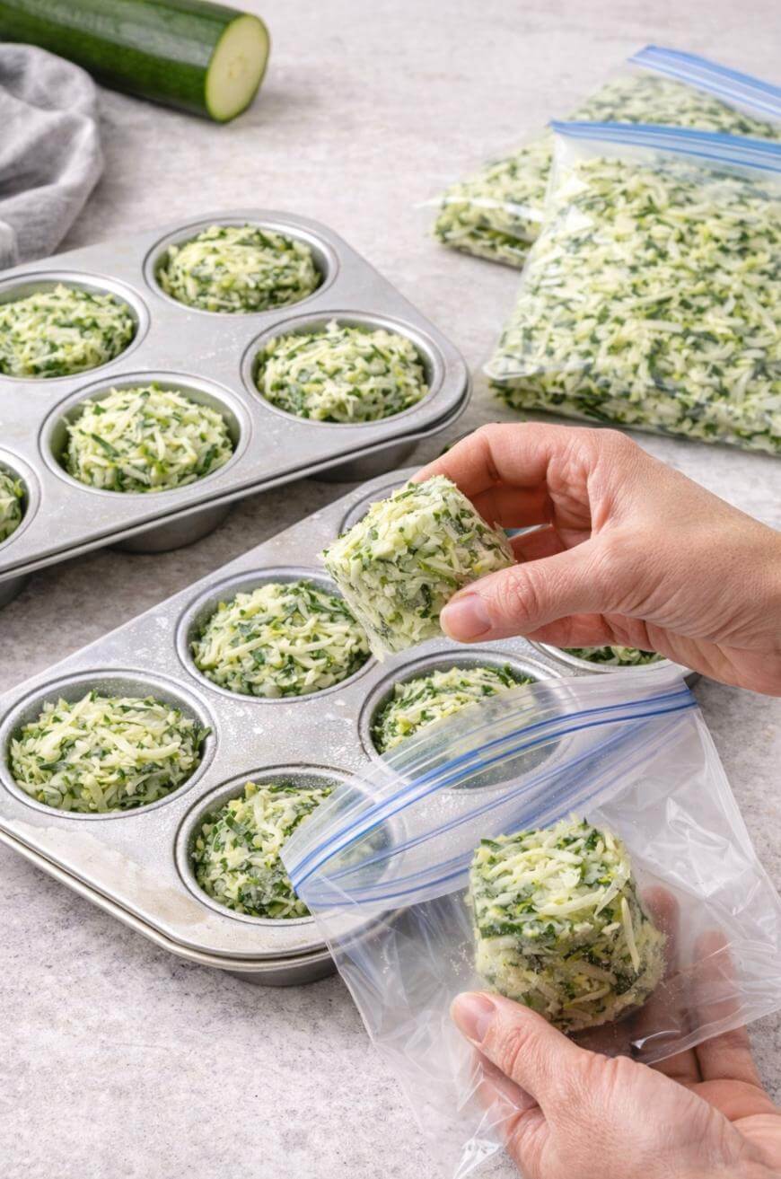 Hands placing a portion of shredded zucchini from a muffin tin into a resealable plastic bag, with more filled muffin tins and bags of shredded zucchini visible on a kitchen counter.