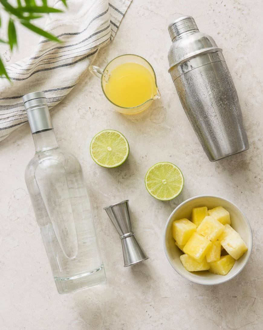 Overhead view of cocktail tools and ingredients: a metal cocktail shaker, a jigger, a clear liquor bottle, a glass of yellow juice, two lime halves, a bowl of pineapple chunks, and a striped towel on a light surface.
