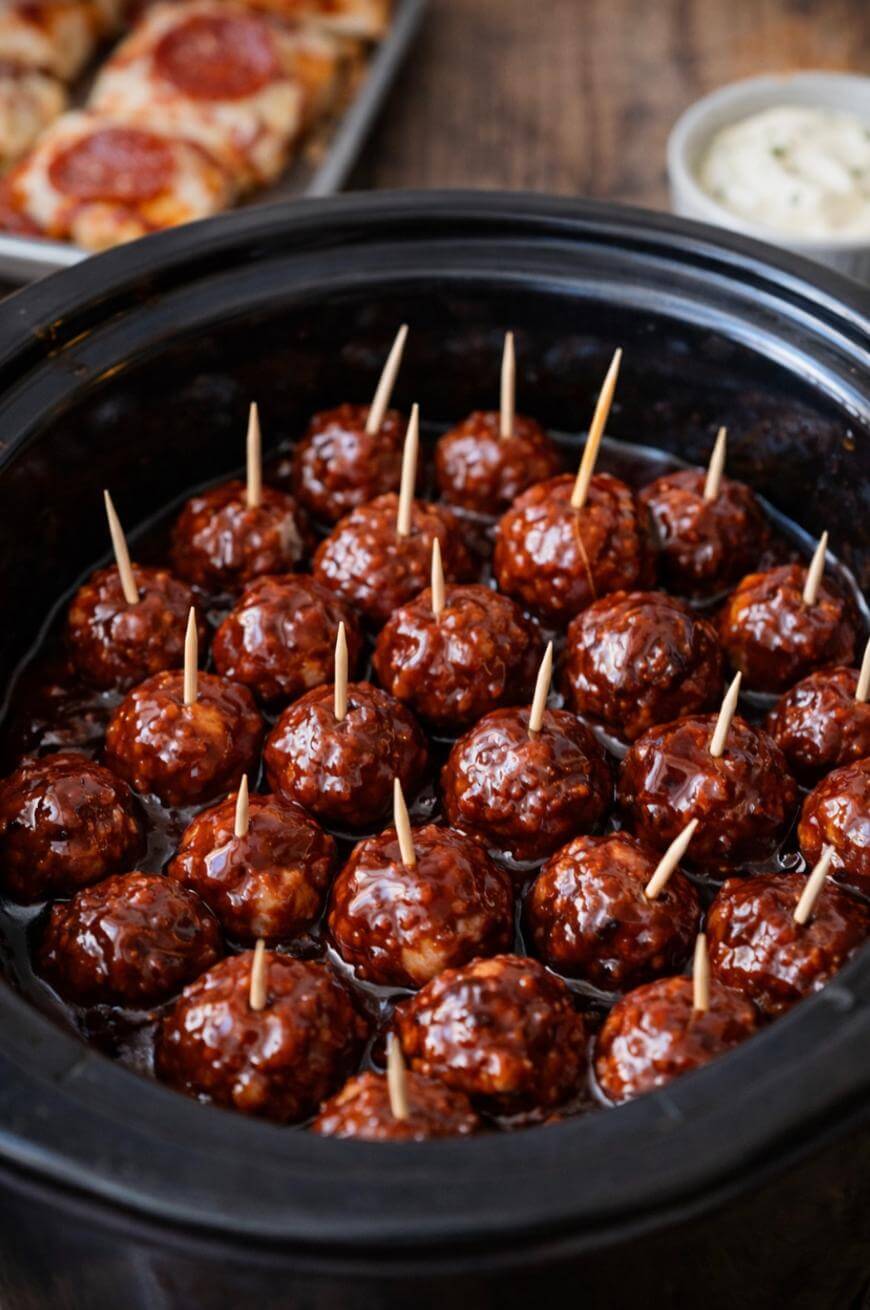 A slow cooker filled with barbecue glazed meatballs, each with a toothpick inserted, sits on a wooden table. In the background, there is a tray of food and a bowl of dipping sauce.