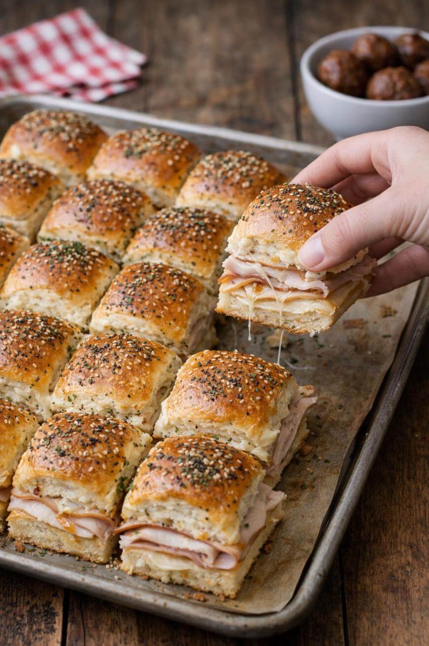A hand lifts a hot turkey and cheese slider topped with sesame and poppy seeds from a baking sheet filled with similar sandwiches. A bowl of meatballs and a napkin are in the background on a wooden table.