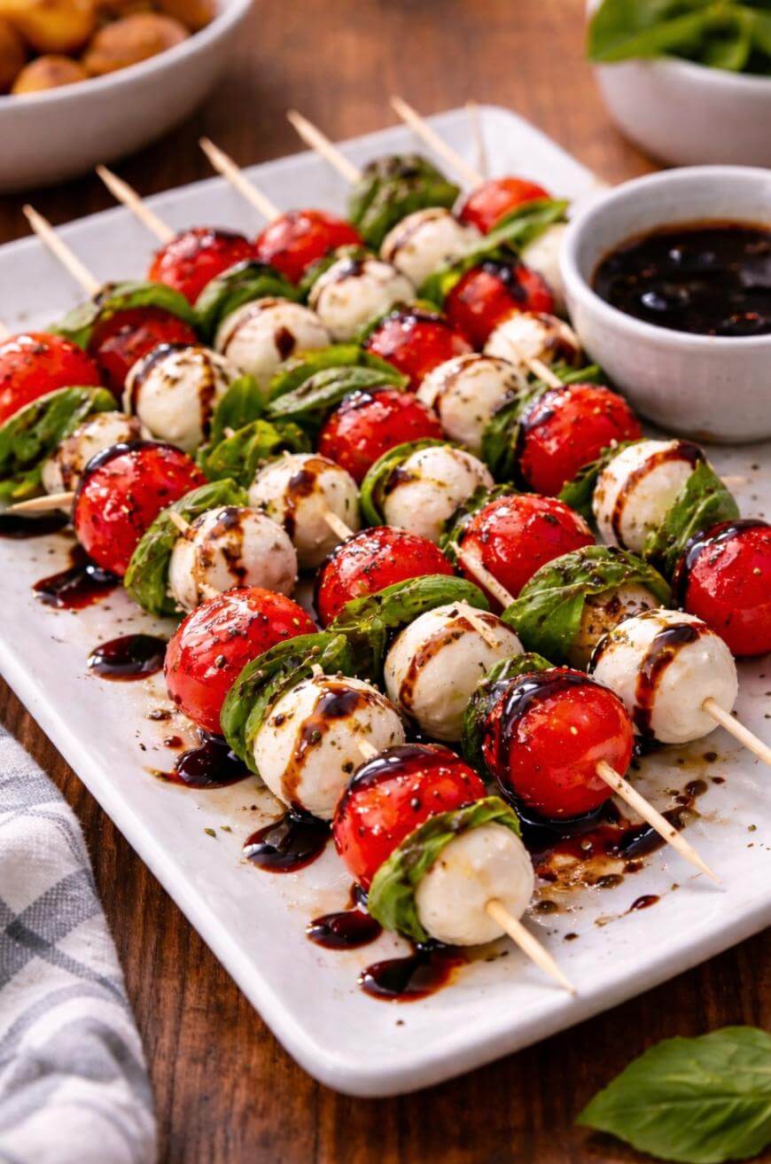 A platter of Caprese skewers with cherry tomatoes, mozzarella balls, and fresh basil leaves, drizzled with balsamic glaze. A bowl of extra balsamic glaze is on the side, and the platter sits on a wooden table.