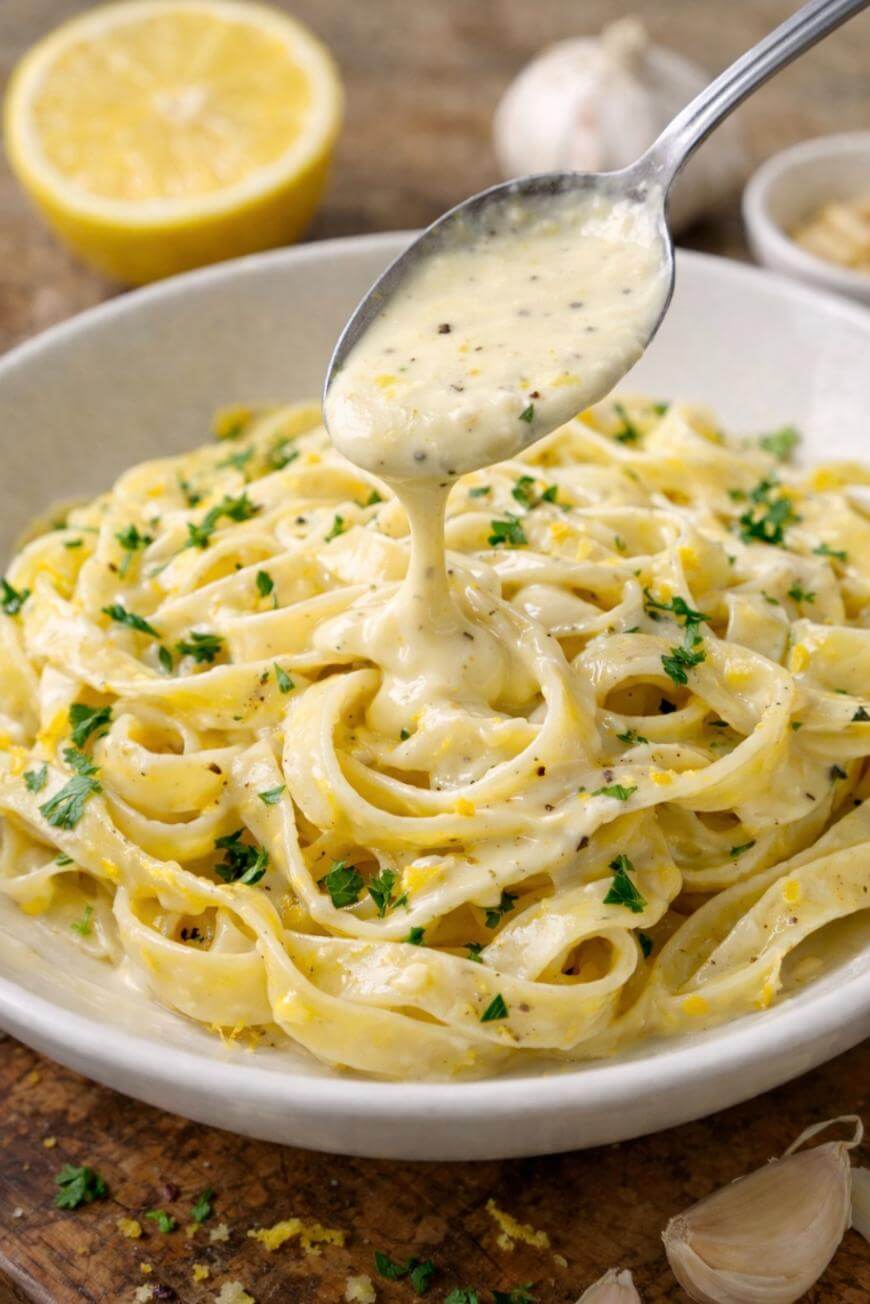 A bowl of fettuccine pasta topped with creamy Alfredo sauce, being drizzled with more sauce from a spoon. The dish is garnished with chopped parsley and surrounded by garlic and a halved lemon.