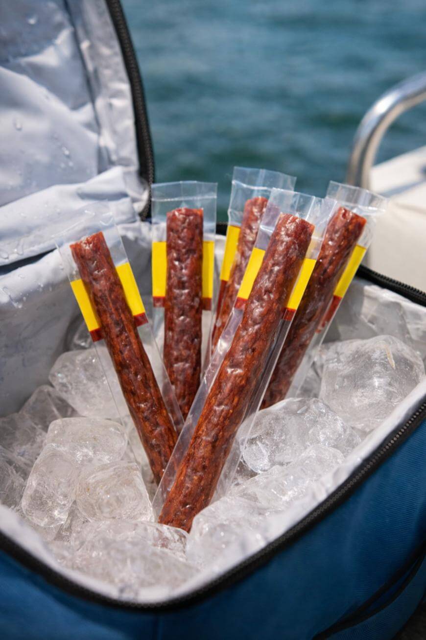 Six packaged beef sticks standing upright in a blue cooler filled with ice, with water visible in the background.