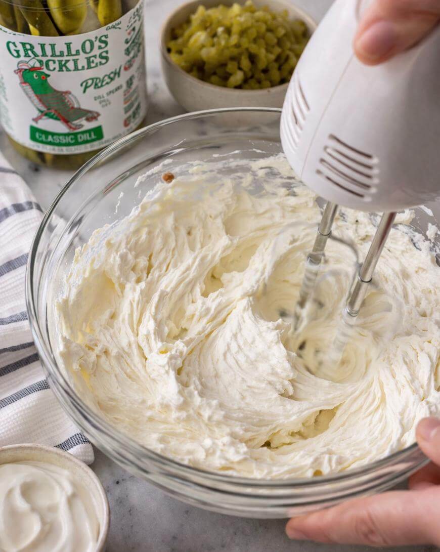 A hand holds a mixer blending creamy white ingredients in a glass bowl. Nearby are a jar of pickles, a bowl of chopped pickles, and a cloth napkin on a marble countertop.
