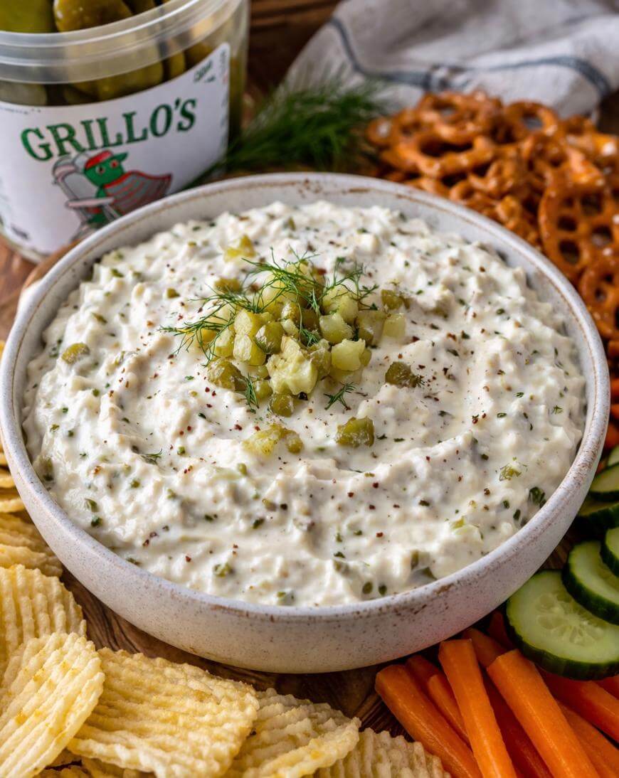 A bowl of creamy dip topped with chopped pickles and dill sits surrounded by potato chips, pretzels, sliced cucumbers, and carrot sticks. A container of Grillo’s pickles is visible in the background.