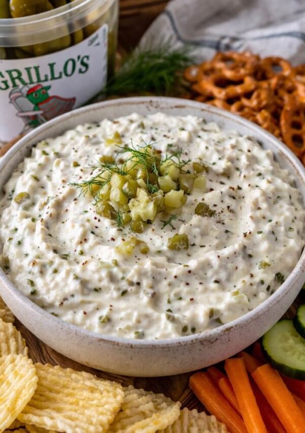 A bowl of creamy dip topped with chopped pickles and dill is surrounded by potato chips, pretzels, carrot sticks, cucumber slices, and a container of pickles in the background.