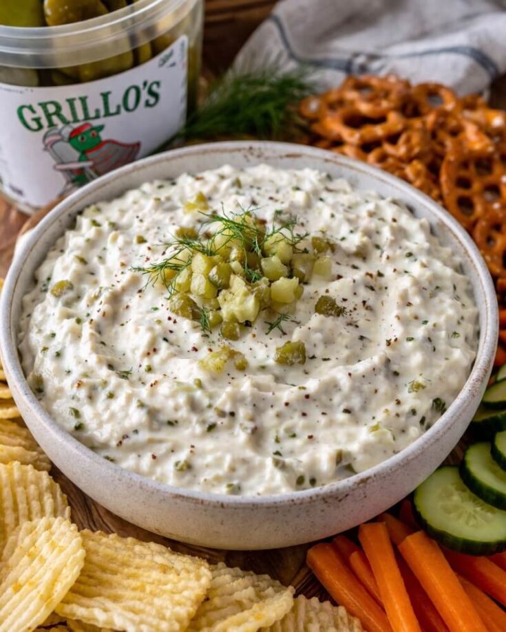 A bowl of creamy dip topped with chopped pickles and dill is surrounded by potato chips, pretzels, carrot sticks, cucumber slices, and a container of pickles in the background.
