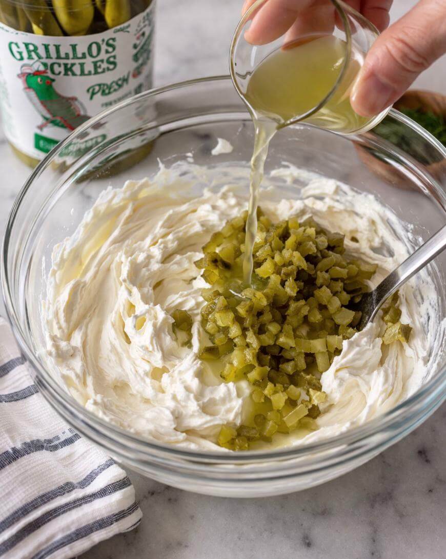 A hand pours pickle juice into a glass bowl of cream cheese and chopped pickles, with a jar of pickles and a striped towel nearby on a marble countertop.