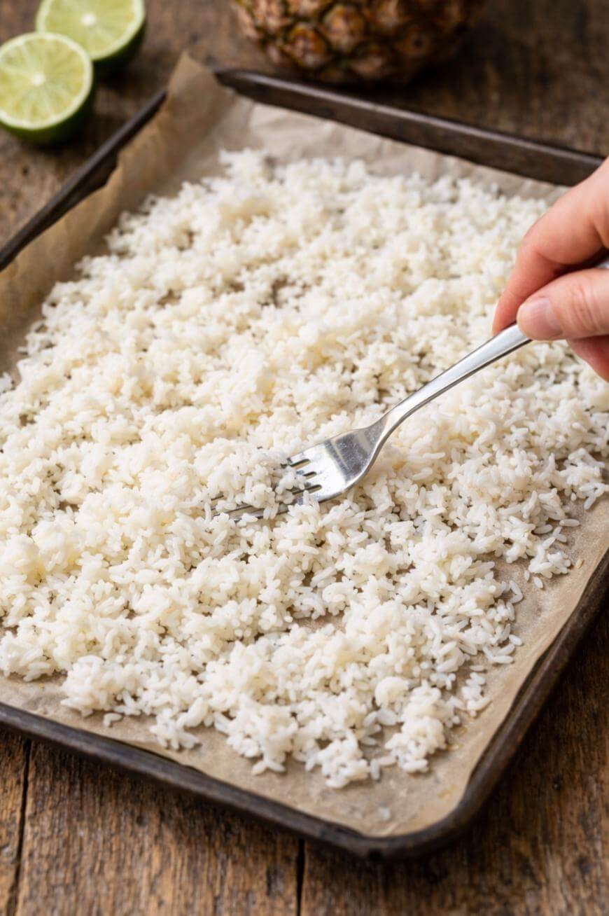 A hand uses a fork to fluff cooked white rice spread on a parchment-lined baking tray. Sliced limes and part of a pineapple are visible in the background on a wooden surface.