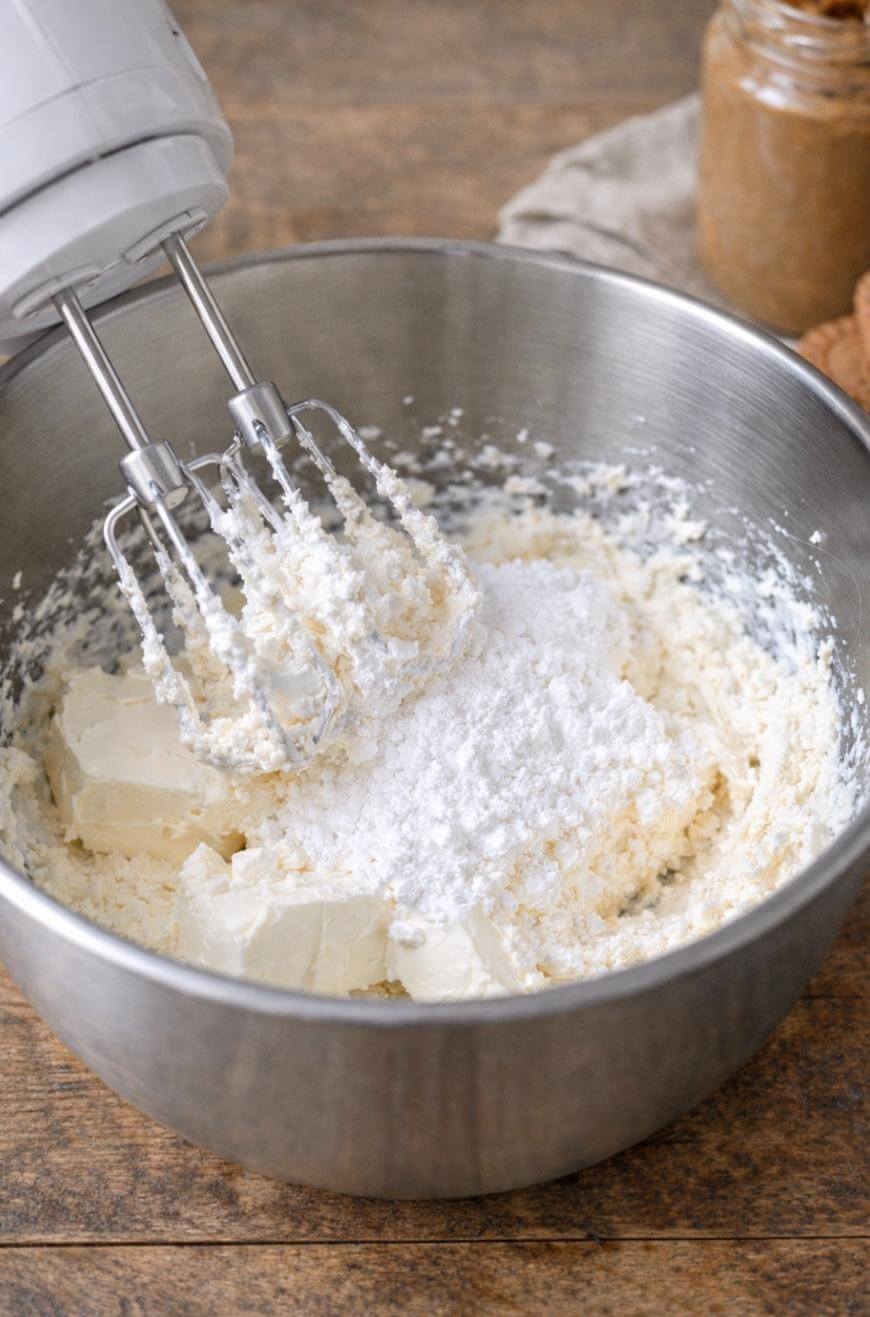 A metal mixing bowl contains creamed butter and powdered sugar being mixed with an electric hand mixer, on a wooden surface with a jar in the background.