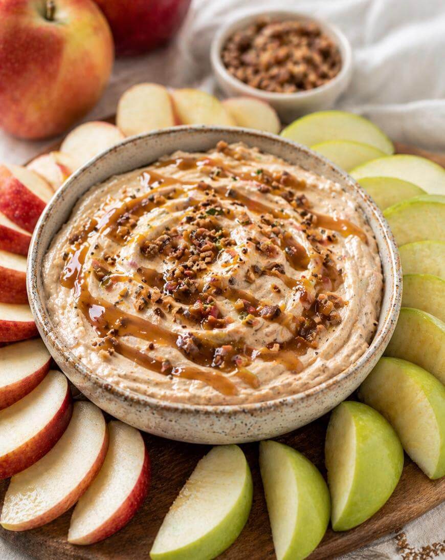 A bowl of creamy dip topped with caramel and chopped nuts is surrounded by slices of red and green apples on a wooden board. In the background, whole apples and a small bowl of nuts are visible.