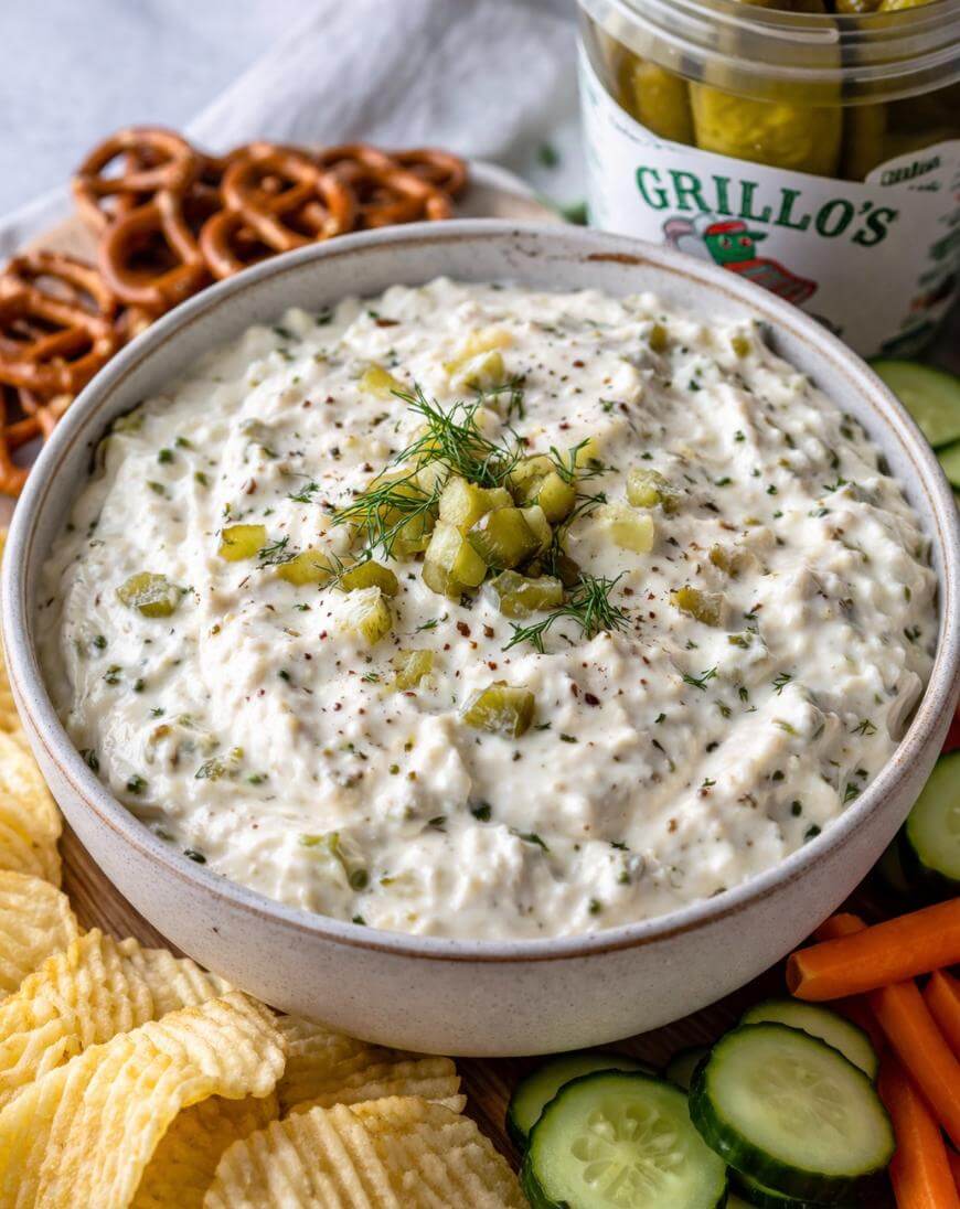 A bowl of creamy dip topped with chopped pickles and dill, surrounded by pretzels, potato chips, cucumber slices, and carrot sticks. An open jar of pickles is in the background.