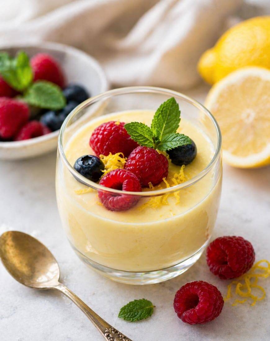 A glass of creamy lemon mousse topped with fresh raspberries, blueberries, lemon zest, and a mint sprig. A gold spoon, extra berries, and lemon halves are nearby on a white surface.
