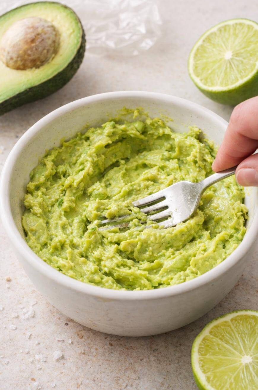A hand mashing avocado with a fork in a white bowl, surrounded by halves of lime and avocado on a light countertop.