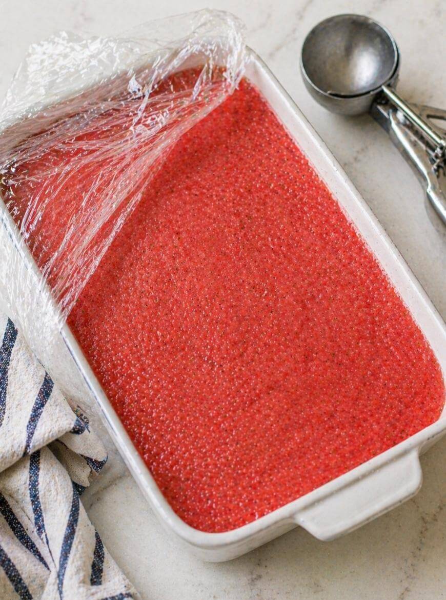 A rectangular white dish filled with bright red granita is partially covered with plastic wrap. An ice cream scoop and a striped kitchen towel are nearby on a light countertop.
